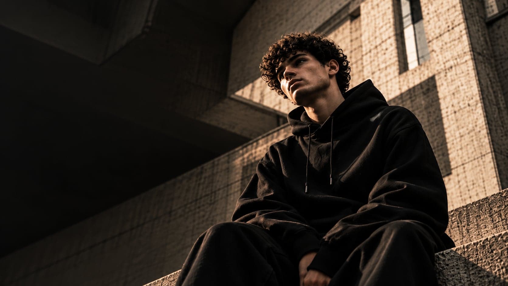 Jeune homme en hoodie noir RASSM assis sur un escalier en béton brut