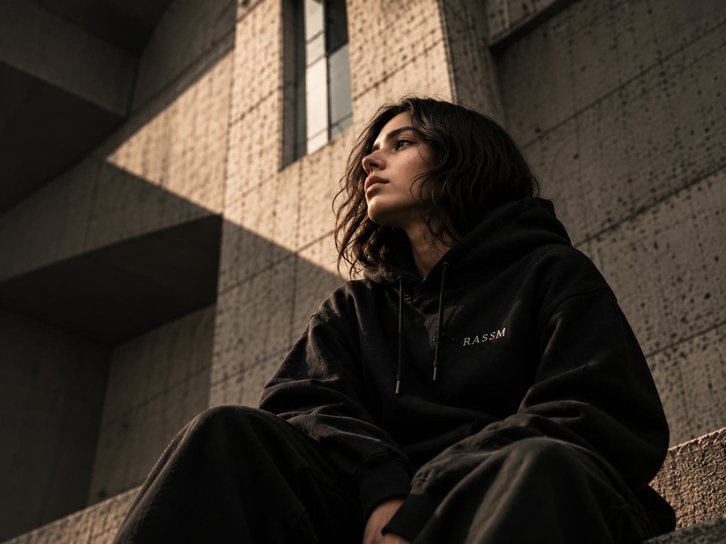 Jeune femme en hoodie noir RASSM, assise devant un mur en béton